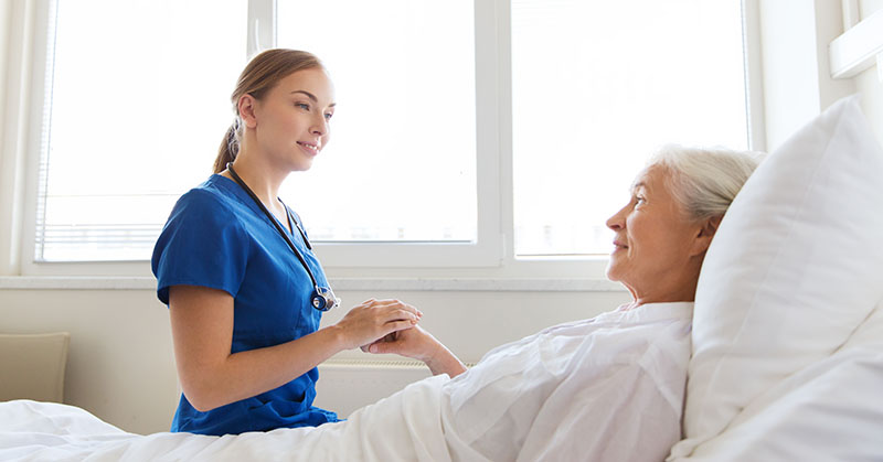 medicine, age, support, health care and people concept - doctor or nurse visiting and cheering senior woman lying in bed at hospital ward